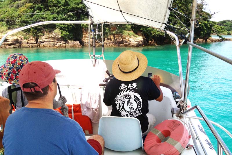 This is the inside of a ferry crossing from Katsumoto Town to Tatsuno Island. The captain, wearing a straw hat and a black T-shirt with the word "Iki" written on it is driving the boat. The sea is a cobalt blue color.