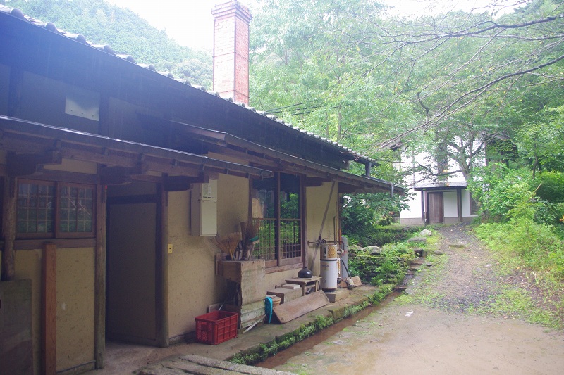 It is the workshop of Ryuta-kiln with the chimney of the kiln for baking ceramic wares and surrounded by green trees.