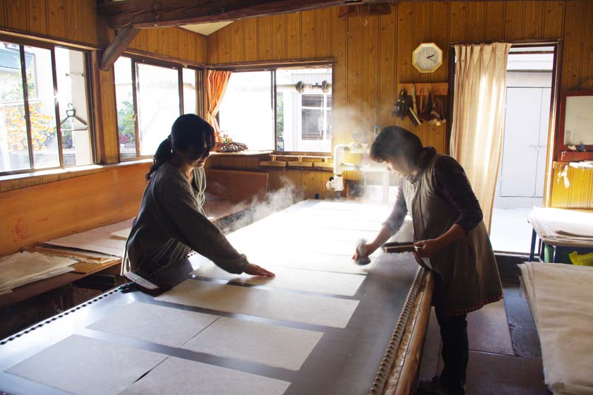 Ini adalah Workshop Matsuo Washi di Yame. Dua wanita sedang mengeringkan washi basah yang baru dibuat di atas meja berpemanas.