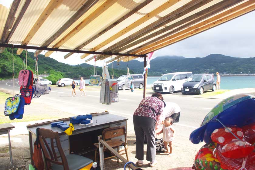 There is a shop for sea bathers near the Hitotsuku beach. There is a store right on the beach that sells floats and flip-flops. A shopkeeper is talking to a customer.