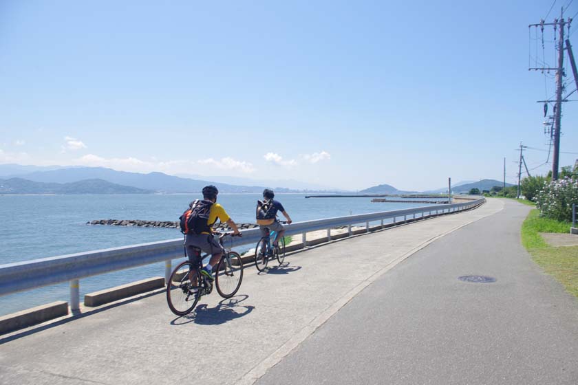 Two people are riding bicycles along a coastal road at the southern tip of Nokonoshima island.